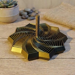 Könnte beinhalten: Eine gold-schwarze geometrische Skulptur mit einem zentralen vertikalen Pfosten. Die Skulptur hat ein geschichtetes, sternförmiges Design mit einer strukturierten Oberfläche. Das Stück wird auf einer Holzoberfläche ausgestellt.