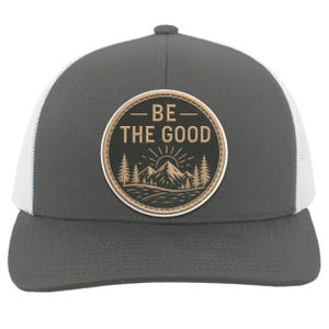 Peut inclure: Une casquette de type trucker gris anthracite et blanc avec un écusson circulaire sur le devant. L'écusson noir à bordure beige porte l'inscription BE THE GOOD au-dessus d'une illustration graphique de montagnes, de sapins et d'un soleil.