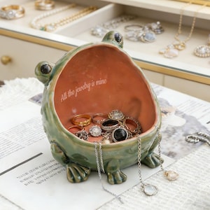 Könnte beinhalten: Eine grüne, froschförmige Schmuckschale aus Keramik, gefüllt mit Ringen und Halsketten. Der Innenraum ist pfirsichfarben mit dem Text "All the jewelry is mine". Die Schale steht auf einer weißen Oberfläche mit anderem Schmuck im Hintergrund.