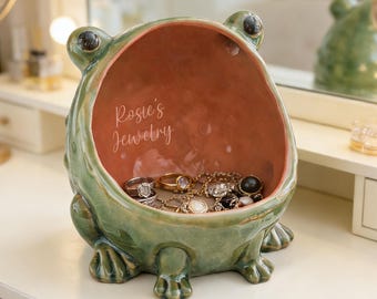 May include: A green ceramic frog-shaped bowl with a wide, open mouth serving as a storage container. The interior is a light terracotta colour with the white text 'Rosie's Jewelry' printed inside. Several metal rings and chains rest at the bottom.