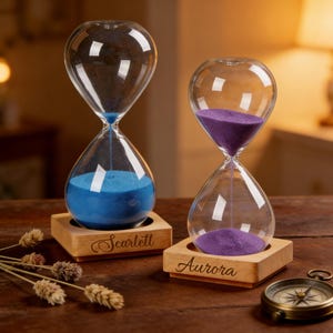 May include: Two glass hourglasses with wooden bases. One contains blue sand, the other purple. The base of the blue hourglass is engraved with the name "Scarlett", while the purple hourglass base says "Aurora". A compass is visible in the foreground.