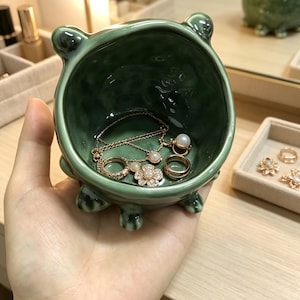 K&ouml;nnte beinhalten: Eine kleine, runde, dunkelgr&uuml;ne Keramikschale mit gl&auml;nzender Oberfl&auml;che und vier kurzen, abgerundeten F&uuml;&szlig;en. Im Inneren befinden sich eine zarte goldfarbene Halskette mit einem kleinen Perlenanh&auml;nger, ein goldener Ring und ein goldener Blumenring mit Perle.