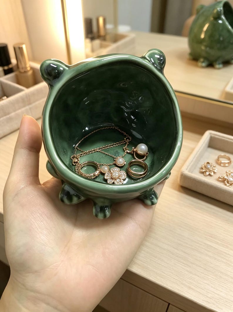 K&ouml;nnte beinhalten: Eine kleine, runde, dunkelgr&uuml;ne Keramikschale mit gl&auml;nzender Oberfl&auml;che und vier kurzen, abgerundeten F&uuml;&szlig;en. Im Inneren befinden sich eine zarte goldfarbene Halskette mit einem kleinen Perlenanh&auml;nger, ein goldener Ring und ein goldener Blumenring mit Perle.