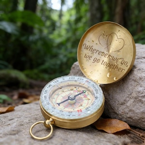 Könnte beinhalten: Ein goldfarbener Kompass mit geöffnetem Deckel, der den Text "Wherever we go, we go together" und zwei Herzformen zeigt. Der Kompass liegt auf einem Stein, mit einem Waldhintergrund.