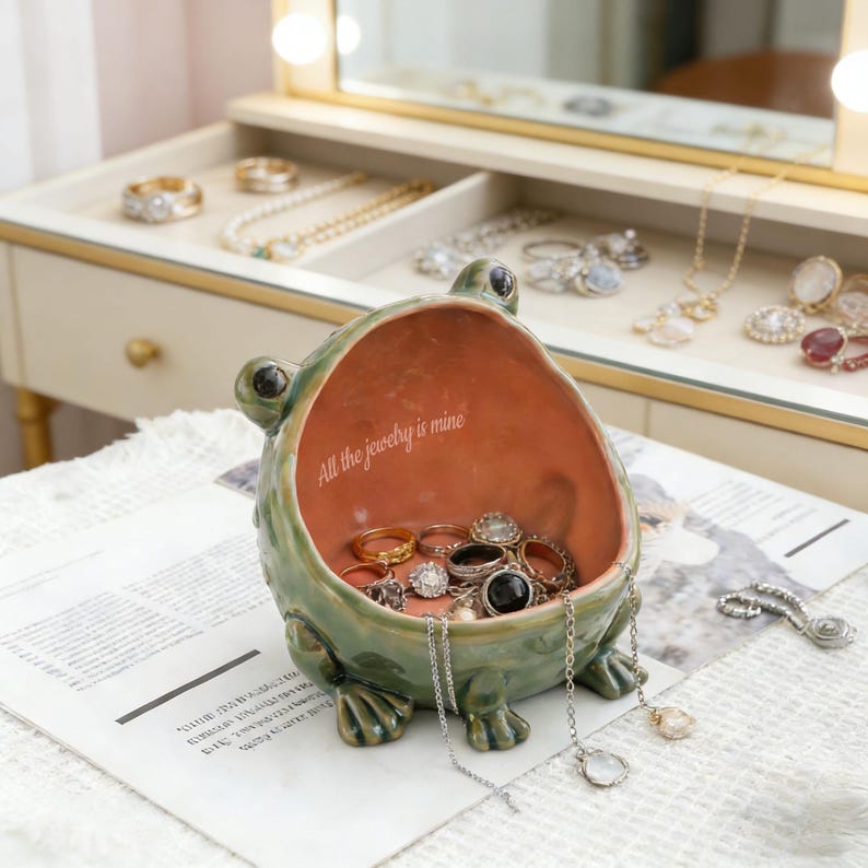 K&ouml;nnte beinhalten: Eine gr&uuml;ne, froschf&ouml;rmige Schmuckschale aus Keramik mit terrakottafarbenem Innenraum. Im Inneren befindet sich der wei&szlig;e Schriftzug "All the jewelry is mine". Die Schale enth&auml;lt verschiedene Metallringe und silberfarbene Ketten am Rand.