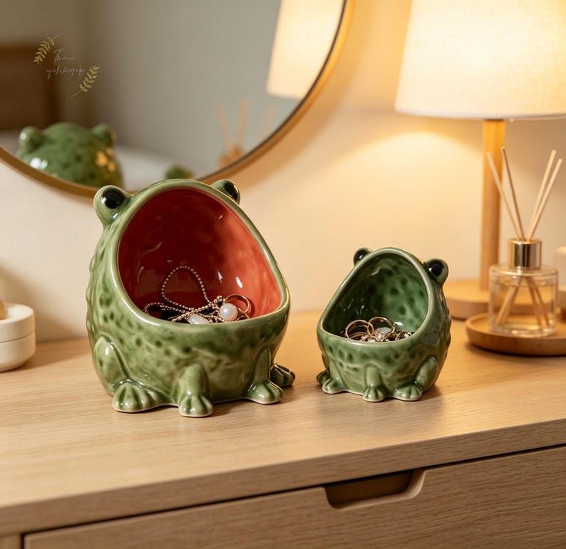K&ouml;nnte beinhalten: Zwei froschf&ouml;rmige Keramikschalen in unterschiedlichen Gr&ouml;&szlig;en. Der gr&ouml;&szlig;ere Frosch hat eine gr&uuml;ne, texturierte Au&szlig;enseite und eine glatte, korallenorangefarbene Innenseite, w&auml;hrend der kleinere Frosch eine gr&uuml;ne Au&szlig;enseite und Innenseite hat.