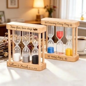May include: Two wooden hourglasses with clear glass tubes and colored sand. One has the text "Time flies, love stays" and the other says "Growing Every Day." The first hourglass has white, gray, and black sand. The second has blue, red, and yellow sand.
