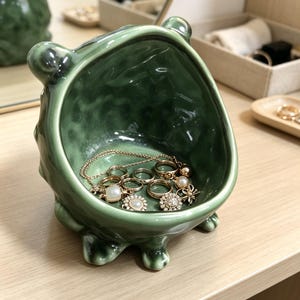 K&ouml;nnte beinhalten: Eine gl&auml;nzende, salbeigr&uuml;ne Keramikschale in Form eines kleinen Wesens mit abgerundeten Ohren und kurzen F&uuml;&szlig;en. Das strukturierte Innere enth&auml;lt eine Sammlung goldfarbener Ringe, eine zarte Kette und kleinen Schmuck mit wei&szlig;en Akzenten.