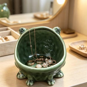 K&ouml;nnte beinhalten: Eine salbeigr&uuml;ne Keramikschale in Froschform mit strukturierter Oberfl&auml;che, vier kleinen F&uuml;&szlig;en und zwei abgerundeten Ohren. Die Schale enth&auml;lt eine goldfarbene Halskette und mehrere Ringe mit wei&szlig;en Steinen und blumenartigen Metalldesigns.