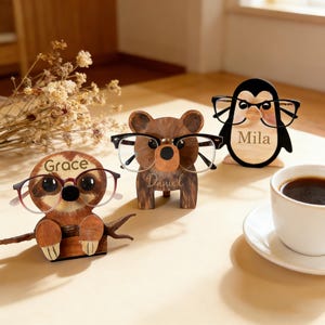 Peut inclure: Trois porte-lunettes en bois en forme de paresseux, d'ours et de pingouin. Chaque support porte un nom gravé : Grace, Daniel et Mila. Le paresseux est brun, l'ours est brun et le pingouin est noir et blanc. Chaque support porte des lunettes.