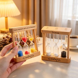 May include: Two wooden hourglasses with glass tubes filled with colored sand. One has blue, red, and yellow sand, the other has white and black sand. Text on the wood reads "Every moment with you is timeless" and "Victoria".
