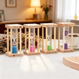 May include: Four wooden hourglasses with coloured sand: blue, pink, green, and purple. Each hourglass has a name engraved on it: Ethan, Olivia, William & Hannah. The hourglasses are on a white surface.