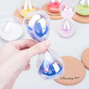 Pode incluir: Ampulheta de vidro iridescente com areia azul, segurada por uma mão. Outras ampulhetas com areia de cores diferentes estão no fundo. Também são visíveis bases de cortiça com o texto "Brushing time".