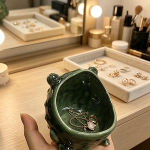 K&ouml;nnte beinhalten: Eine kleine, dunkelgr&uuml;ne Keramikschale f&uuml;r Schmuck mit einer unebenen, strukturierten Oberfl&auml;che und vier kurzen Beinen. Der sch&uuml;sself&ouml;rmige Beh&auml;lter hat eine gl&auml;nzende Oberfl&auml;che und enth&auml;lt eine zarte goldfarbene Kette mit Anh&auml;nger und einen Ring.
