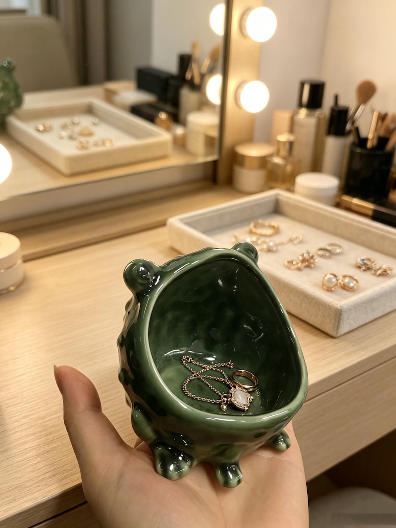 K&ouml;nnte beinhalten: Eine kleine, dunkelgr&uuml;ne Keramikschale f&uuml;r Schmuck mit einer unebenen, strukturierten Oberfl&auml;che und vier kurzen Beinen. Der sch&uuml;sself&ouml;rmige Beh&auml;lter hat eine gl&auml;nzende Oberfl&auml;che und enth&auml;lt eine zarte goldfarbene Kette mit Anh&auml;nger und einen Ring.