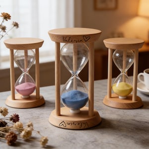 Peut inclure: Trois sabliers en bois avec des ampoules en verre transparent et du sable coloré : rose, bleu et jaune. Chaque sablier a une base et un dessus en bois, avec le nom "Valeria" et un cœur gravé.