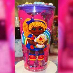 May include: A pink plastic tumbler with a purple lid and a cartoon doll design. The doll has blonde hair, a blue bow, and is holding a stuffed animal. The tumbler has a rainbow design and is filled with liquid and small candies.