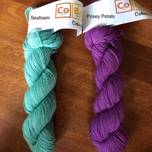 May include: Two skeins of yarn, one in a seafoam green color and the other in a deep purple color. The yarn is labeled "Cobblestone" and the colors are "Seafoam" and "Posey Petals".