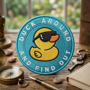 May include: A round, teal embroidered patch with the text "DUCK AROUND AND FIND OUT" encircling a yellow duck wearing sunglasses. The duck has an orange beak and is centered on the patch. The patch is on a wooden surface with a compass, pen, and other items.
