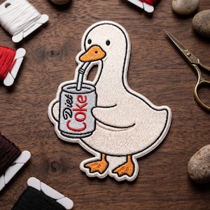 May include: Embroidered patch featuring a white goose drinking from a Diet Coke can with a straw. The goose has an orange beak and feet. The patch is surrounded by sewing supplies, including thread spools and scissors, on a wooden surface.