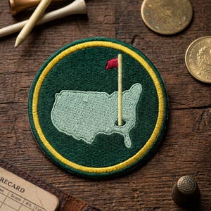May include: A circular embroidered patch featuring a green map of the United States with a golf flag. The patch has a yellow border and a red flag. Golf tees and coins are visible in the background.