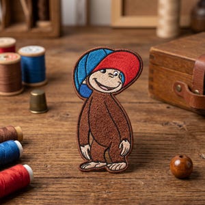 May include: Embroidered patch featuring a cartoon monkey with a red and blue hat. The monkey is brown with a smiling face. The patch is on a wooden surface with spools of thread and a wooden box in the background.