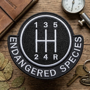 May include: Black and white embroidered patch with the words "ENDANGERED SPECIES" and a gear shift pattern. The patch features the numbers 1, 2, 3, 4, 5, and R. The patch is round with a black background and white stitching.