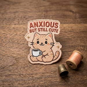 May include: Embroidered patch featuring a cartoon cat holding a coffee cup, with the text "ANXIOUS BUT STILL CUTE". The patch is light brown with darker brown accents. A spool of thread and a thimble are also visible.