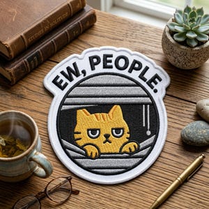 May include: Embroidered patch with the words "EW, PEOPLE" above a grumpy yellow cat peeking through blinds. The patch is white with black and gray details. The cat has a grumpy expression. The patch is on a wooden surface.