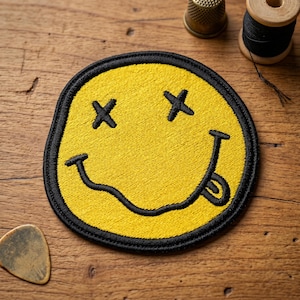 May include: Embroidered yellow patch featuring a smiley face with black cross-eyes and a tongue sticking out. The patch has a black border and is likely designed to be sewn or ironed onto clothing or accessories. The patch is on a wooden surface.