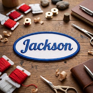 May include: An embroidered patch with the name "Jackson" in blue script lettering on a white oval, framed by a blue border. The patch is surrounded by sewing supplies, including spools of red and white thread, scissors, and a thimble, on a wooden surface.