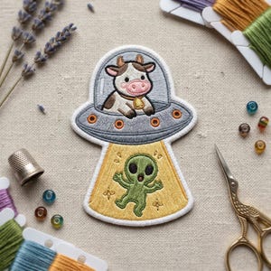 May include: Embroidered patch featuring a cow in a UFO, with an alien below. The cow is brown and white, wearing a gold bell. The UFO is gray with orange lights, and the alien is green. The patch is on a beige background.