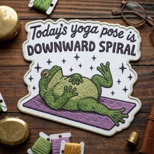 May include: Embroidered patch featuring a green frog in a yoga pose on a purple mat, with the text "Today's yoga pose is DOWNWARD SPIRAL". The patch has a white border with black text and star accents, perfect for clothing or bags.