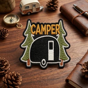 May include: Embroidered patch with the word "CAMPER" in yellow, above a black camper trailer design. Two green and white pine trees flank the camper. The patch is on a wooden surface with other camping-themed items.