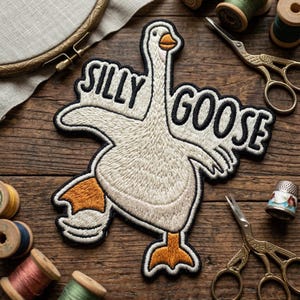 May include: Embroidered patch featuring a white goose with orange feet and beak, outlined in black. The words "SILLY GOOSE" are embroidered above the goose. The patch is on a wooden surface with sewing supplies.