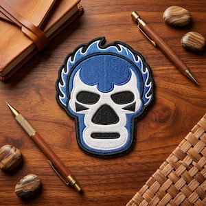 May include: Embroidered patch featuring a blue and white luchador mask design with a black outline. The patch is surrounded by a blue flame-like border. The image also includes two wooden pens, two decorative stones, a leather-bound notebook, and a woven mat.
