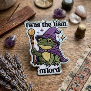 May include: Embroidered patch featuring a green frog dressed as a wizard, wearing a purple hat and cape, holding a staff. The patch has the text "twas the 'tism" and "m'lord". The patch is on a wooden surface.
