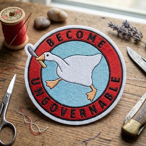 May include: A circular embroidered patch featuring a white goose with orange feet holding a silver knife in its beak. The goose is centered on a light blue background surrounded by a red border with the black text BECOME UNGOVERNABLE.