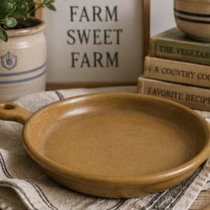 May include: A rustic, tan-colored ceramic skillet with a handle rests on a striped linen cloth. A wooden spoon and a sign that reads "FARM SWEET FARM" are also visible. A stack of vintage cookbooks and a stoneware pot with greenery complete the farmhouse aesthetic.