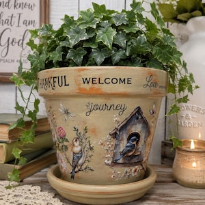 May include: A beige ceramic plant pot with a matching saucer, featuring a green ivy plant. The pot is decorated with images of birds, a birdhouse, and flowers, along with the printed text "THANKFUL", "WELCOME", "Love", and "the journey".