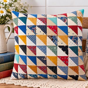 May include: A square throw pillow featuring a patchwork design composed of numerous small triangles in various colors and patterns, including red, blue, yellow, green, and cream, arranged in a grid of half-square triangle blocks.