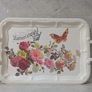 May include: A white rectangular serving tray with a floral and butterfly design. The tray features a decorative border and handles. The design includes roses in shades of pink, yellow, and red, along with a brown butterfly and the text "Herboristerie".