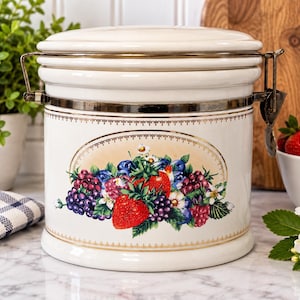 May include: A cream-colored ceramic canister with a metal latch lid. The front features an oval illustration of strawberries, blueberries, and raspberries with green leaves, framed by a delicate gold decorative border.