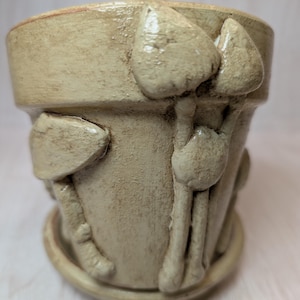 May include: A beige ceramic plant pot with a matching saucer, featuring raised, three-dimensional mushroom sculptures applied to the exterior surface. The pot has a textured, matte finish with subtle brown shading in the crevices.