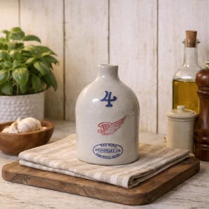 May include: A vintage stoneware jug with a blue "4" and a red wing design. The jug is from Red Wing Stoneware Co. and sits on a wooden cutting board with a striped cloth. A rustic kitchen scene.