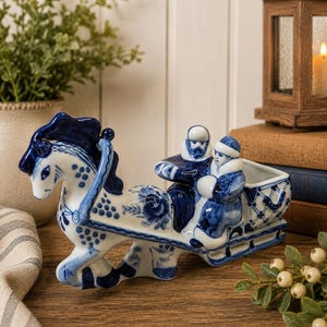 May include: A white ceramic figurine featuring a horse pulling a sleigh with two figures inside. The piece is decorated with intricate blue floral and lattice patterns. The horse is depicted in motion, and the sleigh has a hollow compartment at the back.