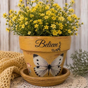 May include: A yellow terracotta flower pot with a matching saucer, featuring the word Believe in black script and a large white butterfly with black spots on the front. The pot is filled with a dense arrangement of small, bright yellow flowers.