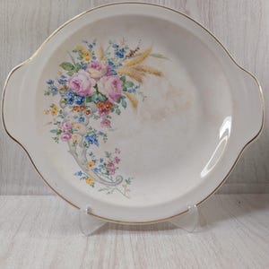 May include: A round white ceramic serving platter with integrated side handles and a gold-trimmed rim. The center features a colorful floral cornucopia design with pink roses, blue forget-me-nots, yellow blossoms, and golden wheat stalks.