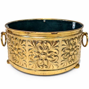 May include: A round brass planter featuring an embossed floral pattern on the exterior, two side ring handles, and three small feet. The interior is dark-colored and the metal surface shows a polished gold-tone finish with some patina.