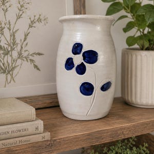 May include: A white ceramic vase with a blue floral design. The vase is cylindrical with a slightly flared top. The flower has five petals and a stem. The vase is sitting on a wooden shelf with books and a framed picture.